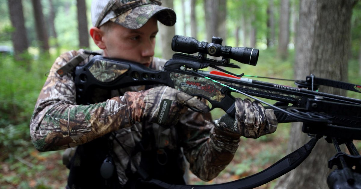 A shooter in camouflage aiming a scoped crossbow in a wooded area