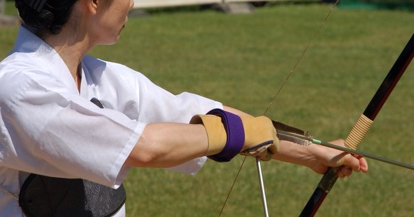 Traditional archer wearing a purple arm guard on the bow arm while drawing a long bow