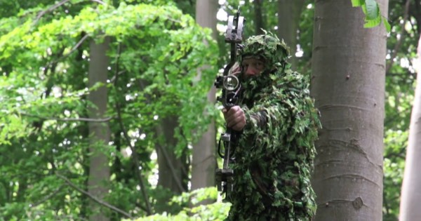 A bowhunter in leafy camouflage aiming a compound bow from among trees