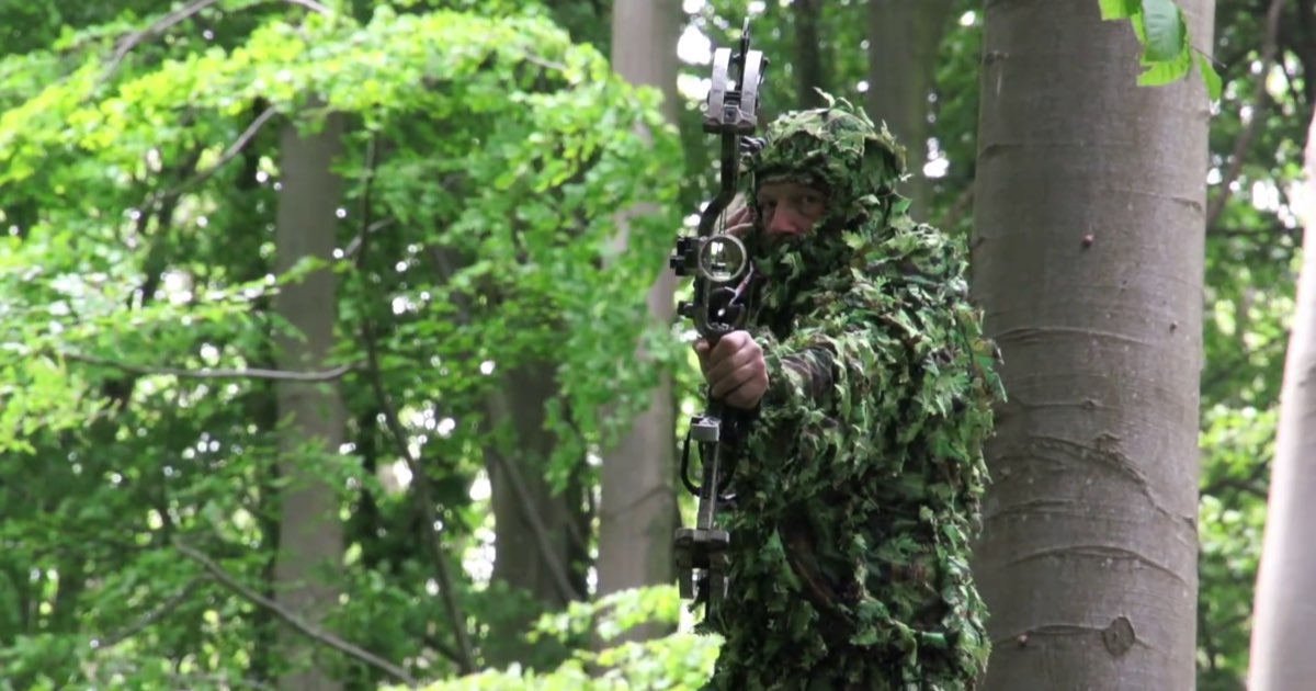 A bowhunter in leafy camouflage aiming a compound bow from among trees