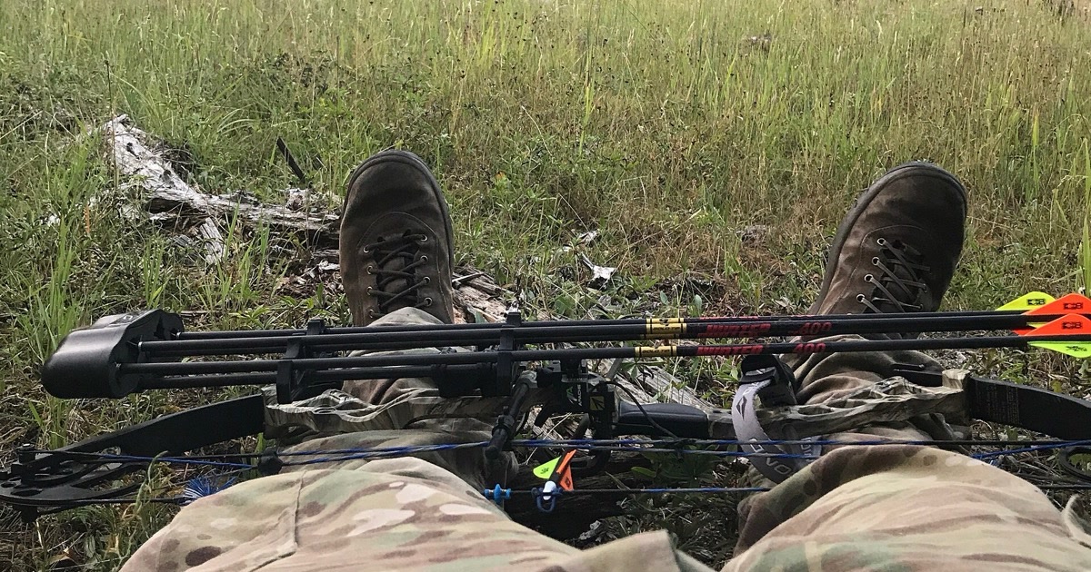 A compound hunting bow with an attached quiver of arrows resting across a bowhunter's lap at the edge of a mountain meadow