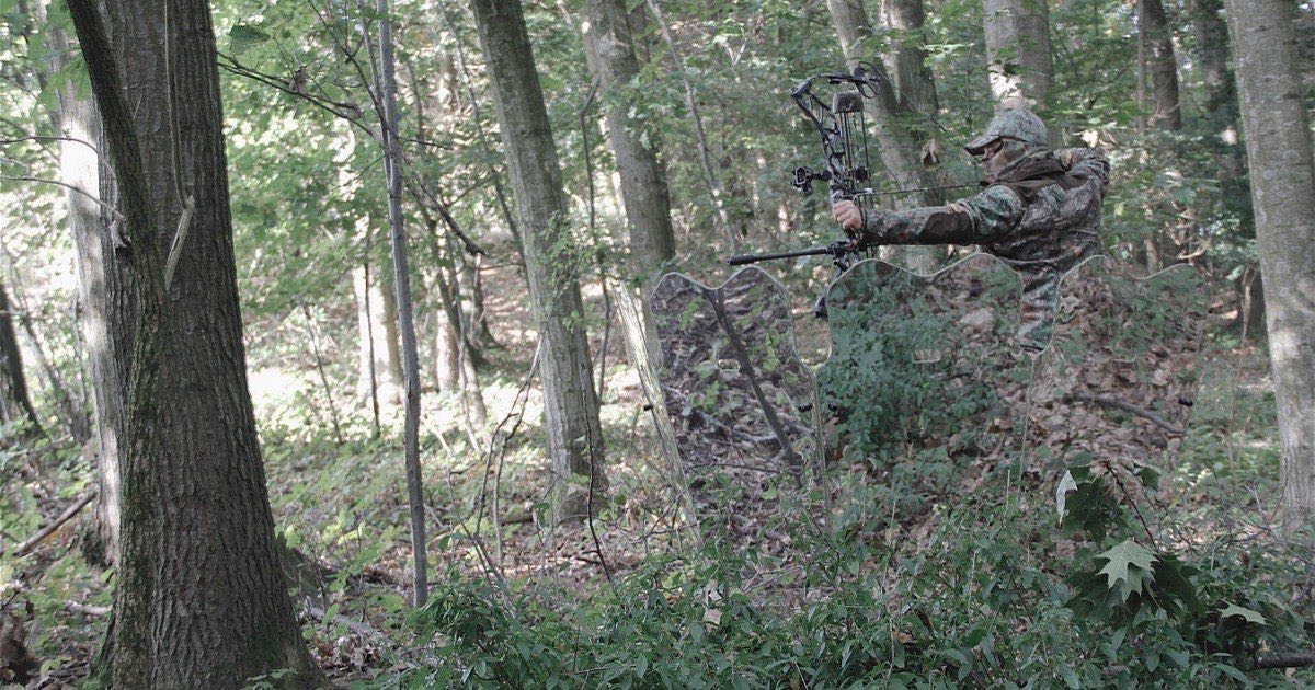 Bowhunter in camouflage at full draw with a compound bow among the trees of a deciduous forest
