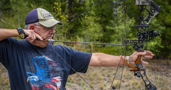 A bowhunter at full draw with a camouflage compound hunting bow fitted with a pin sight, sighting in at an outdoor archery range among pine trees