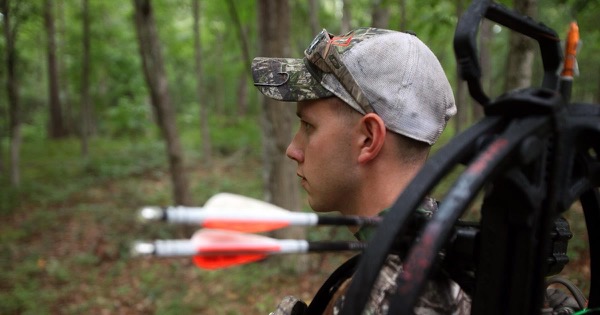 Bowhunter in a camouflage cap walking through summer woodland with a compound bow and arrows slung on his back