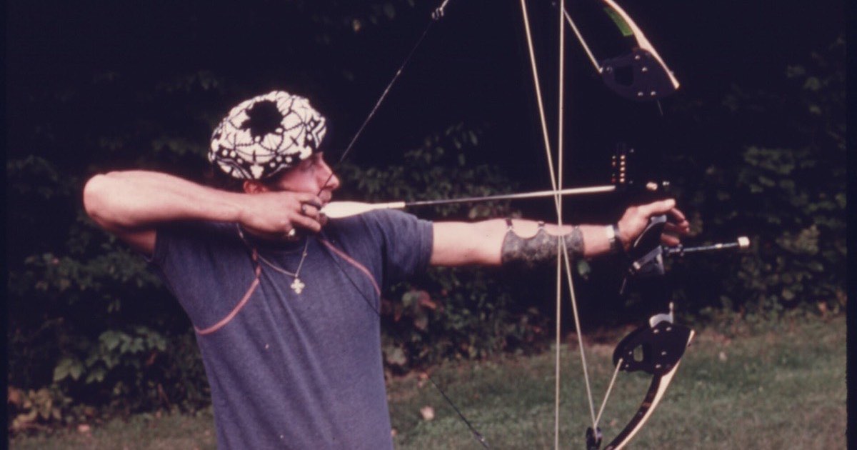 Bowhunter in 1975 drawing an early compound bow with a pin sight while sighting in on a field course