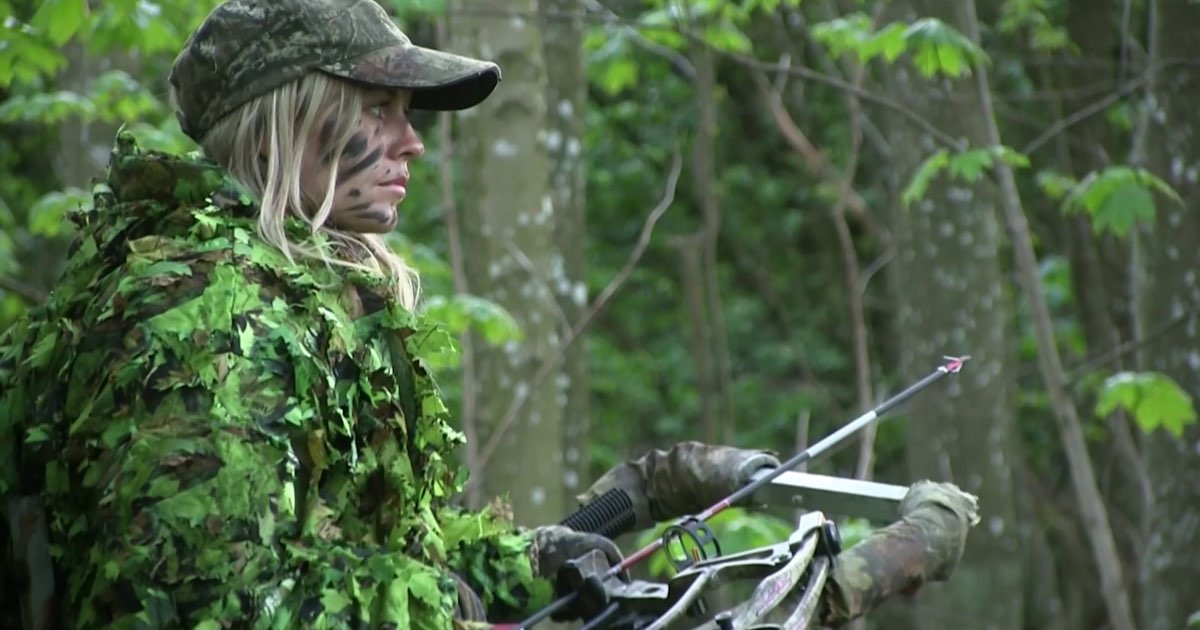 A bowhunter in camouflage holding a compound bow while moving through green woodland
