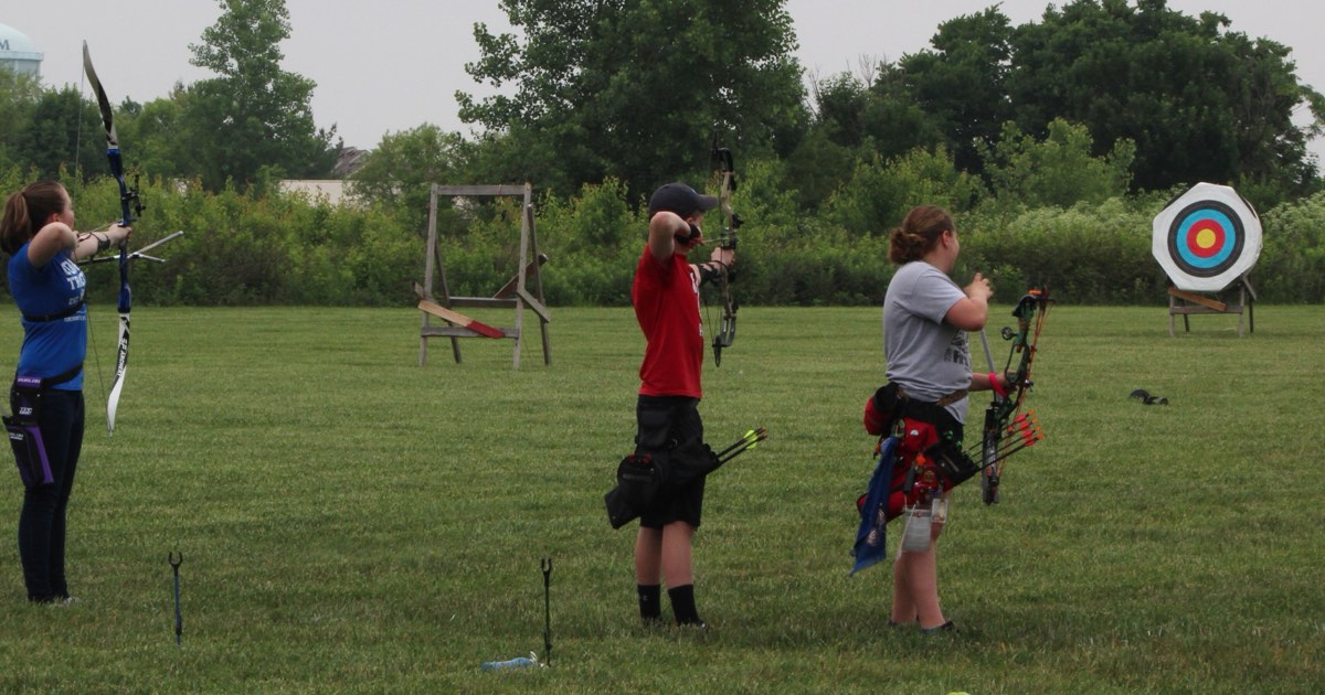 Three archers shooting side by side at an outdoor club range with target faces in the distance