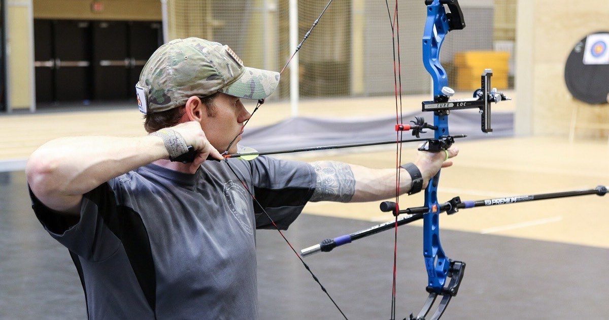 Archer in a cap at full draw with a blue compound bow on an indoor range