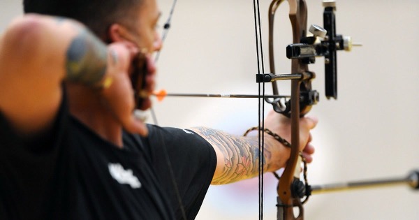 Compound archer at full draw seen from behind the shoulder, with a release aid at the face, a sight on the riser and the bow arm extended