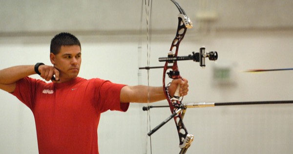A compound archer at full draw with a pin sight and stabiliser on an indoor range