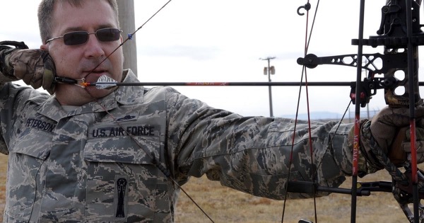 Archer in a camouflage uniform at full draw with a compound bow, the arrow sitting on a raised drop-away rest at the riser