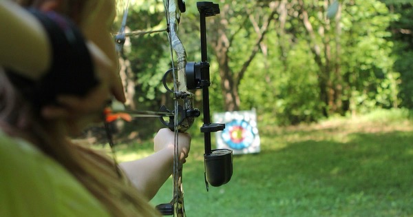 Over-the-shoulder view of a long-haired archer at full draw with a camouflage compound bow, aiming at a target in a backyard clearing
