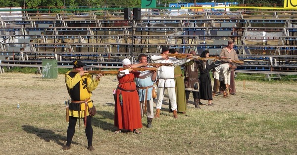 A line of people in medieval costume aiming wooden crossbows on an open shooting field at a festival