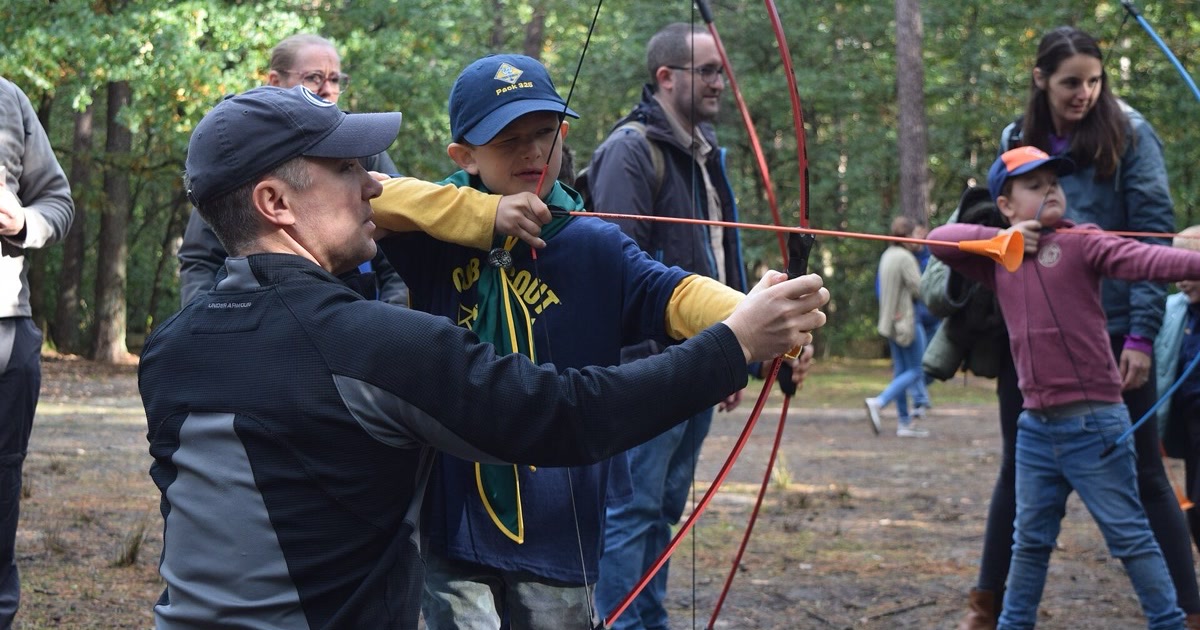 A parent helps a Cub Scout draw a light recurve bow with a soft-tipped arrow at a camporee archery range