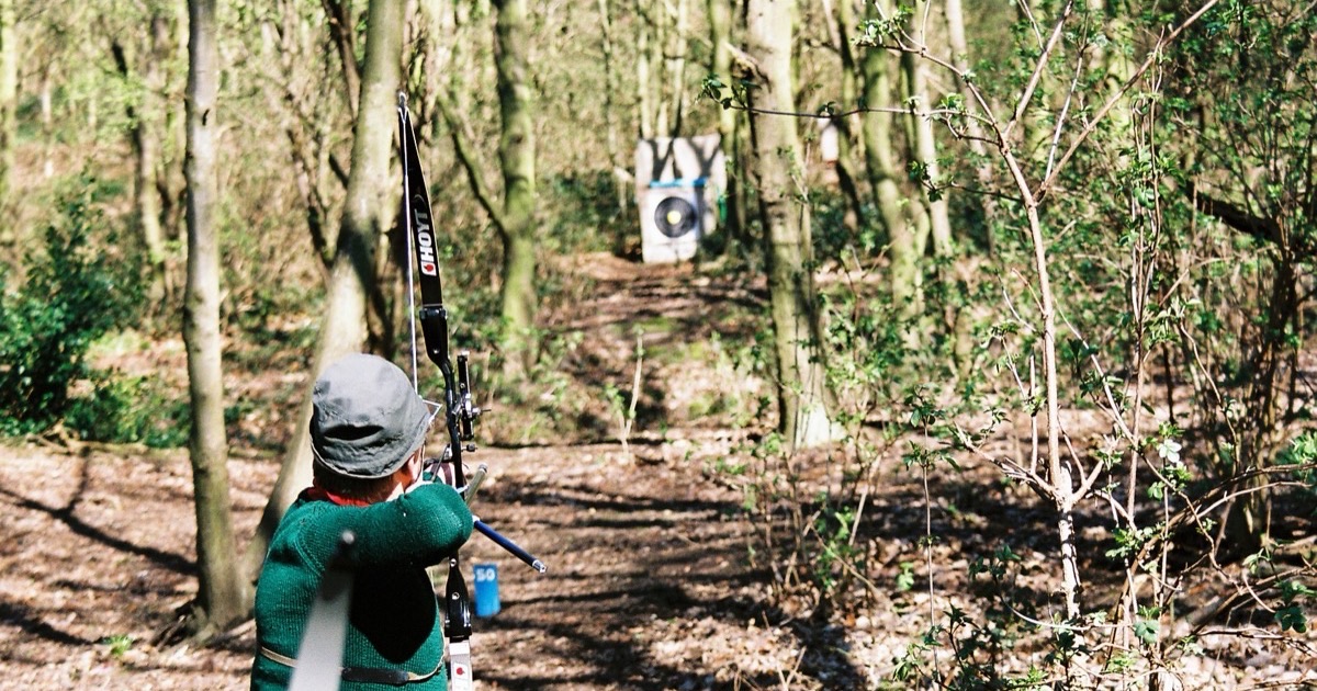 Field archer at full draw with a recurve bow, aiming along a woodland lane at a distant target