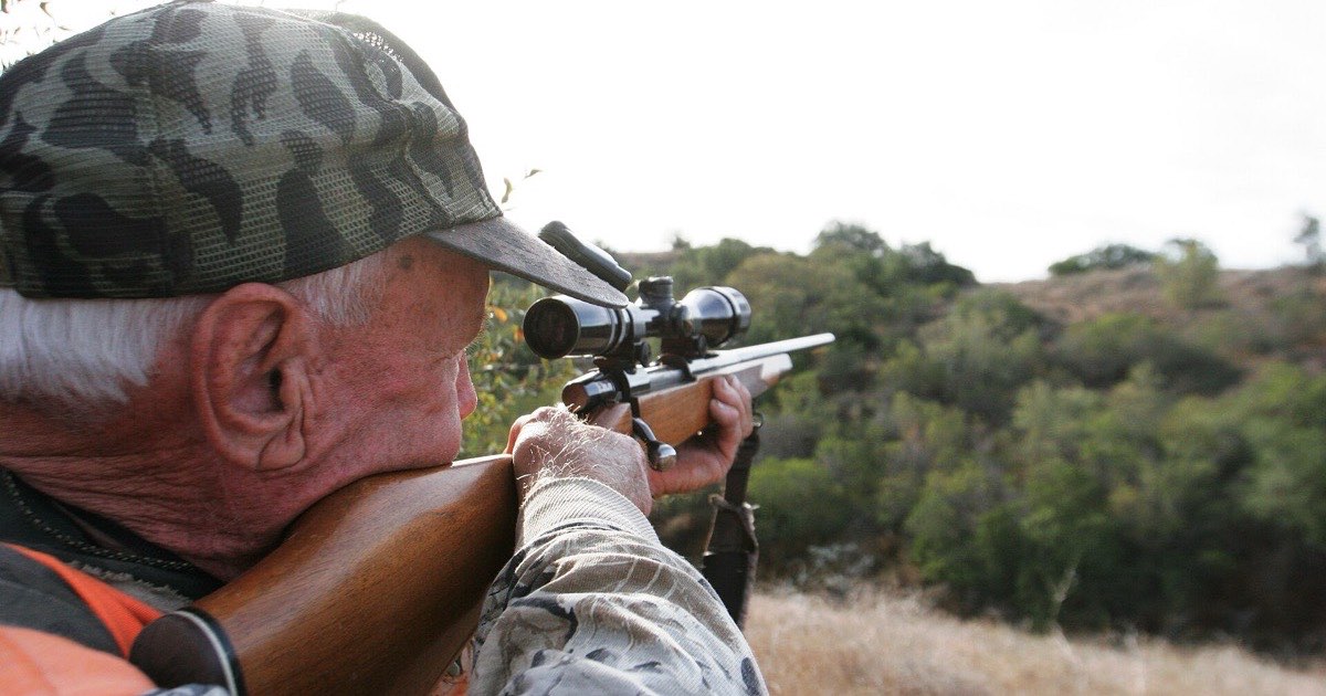Older hunter in camouflage aiming a scoped hunting rifle across open, brushy country