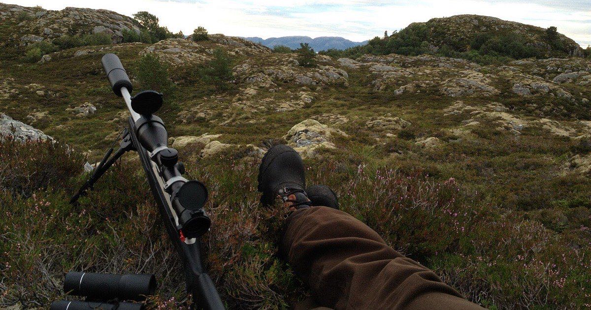 Hunting rifle on a bipod beside a resting hunter on a rocky hillside, with binoculars nearby