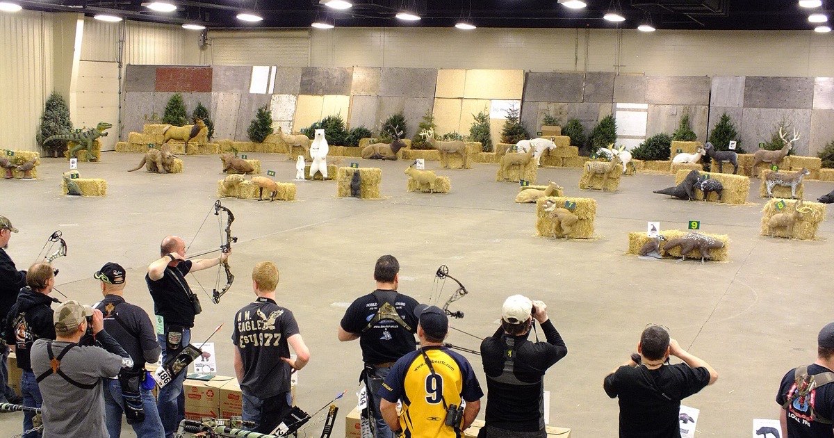 Indoor 3D archery tournament with compound archers at the shooting line and foam animal targets set on hay bales across the hall