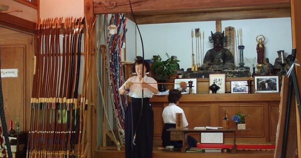 A kyudo archer holding a tall yumi in a temple dojo beside a rack of Japanese bows at Engaku-ji, Kamakura