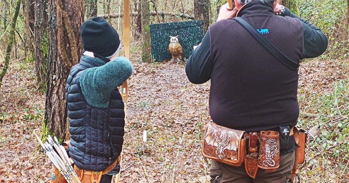 Two archers on a wooded 3D course, one drawing a longbow at a target among the trees