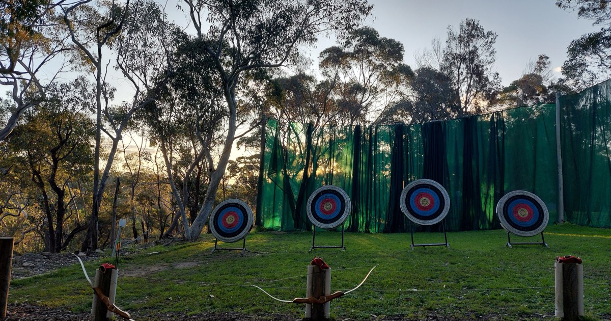 Recurve bows resting on wooden stands at an outdoor range with four targets at dusk