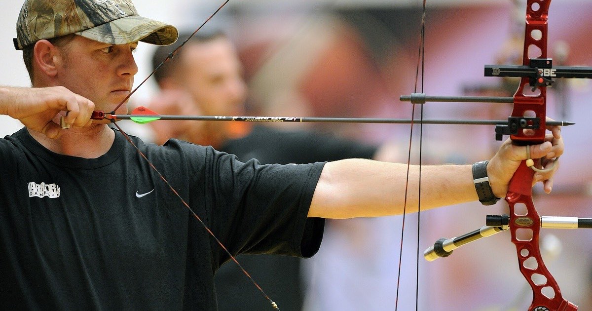 Archer at full draw with a red compound bow fitted with a sight and stabilizer