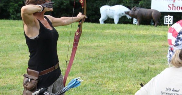 A woman drawing a longbow at an outdoor traditional archery event with 3D targets behind