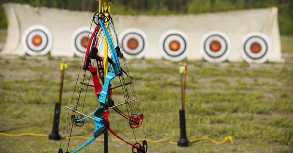 Brightly coloured youth compound bows hanging on a stand in front of a row of target faces at an outdoor range