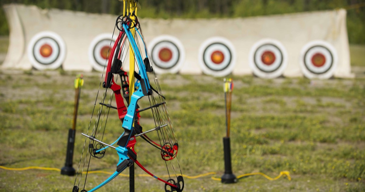 Brightly coloured youth compound bows hanging on a stand in front of a row of target faces at an outdoor range